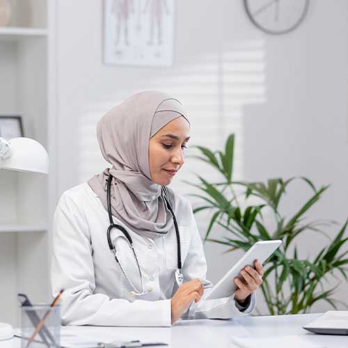 A professional Muslim female doctor in a hijab sits in her office, using a digital tablet. The modern clinic setting portrays a blend of technology and healthcare.