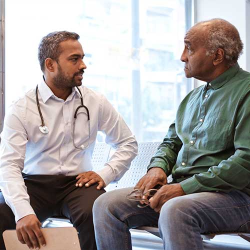 Doctor talking to senior patient sitting in waiting room. Serious man is staring at healthcare worker at hospital. They are against window.