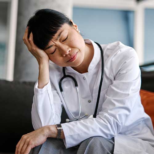 Tired Japanese doctor is sitting on a sofa, her head in her hand, eyes closed, taking a moment to rest after a long and demanding shift at the hospital