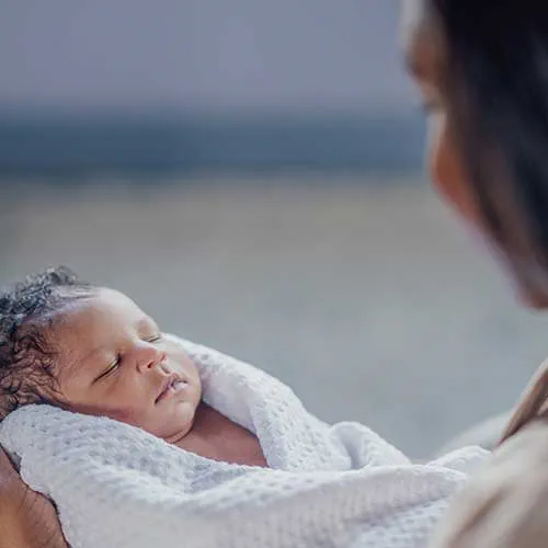 A newborn baby boy, of African decent, lays swaddled in his Mothers arms as he rests at home. The Infant has is eyes closed and a serene expression on his face.