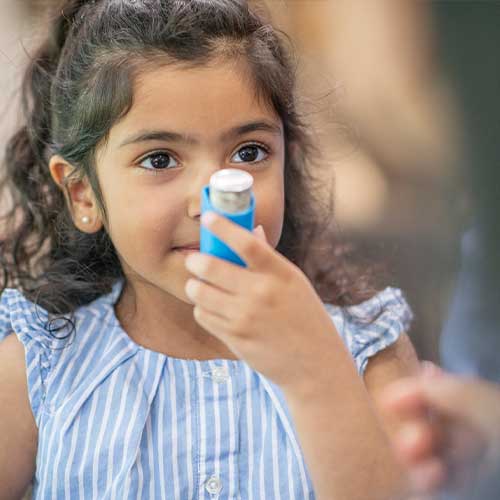 A young girl of Hispanic ethnicity is holding an asthma inhaler close to her mouth to demonstrate how she uses it.