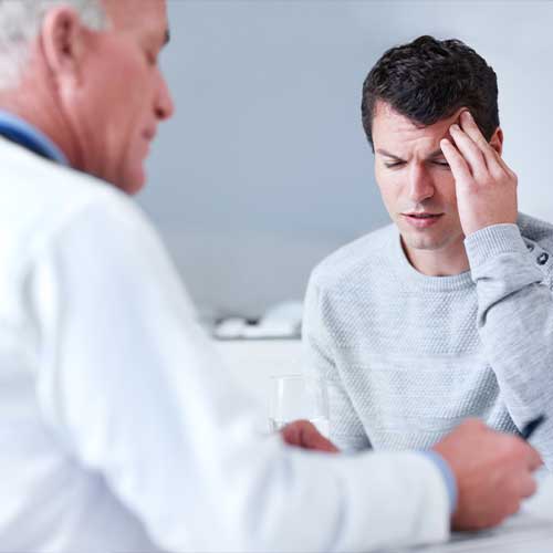 Young man rubbing his forehead in agony while his doctor is writing out a prescription