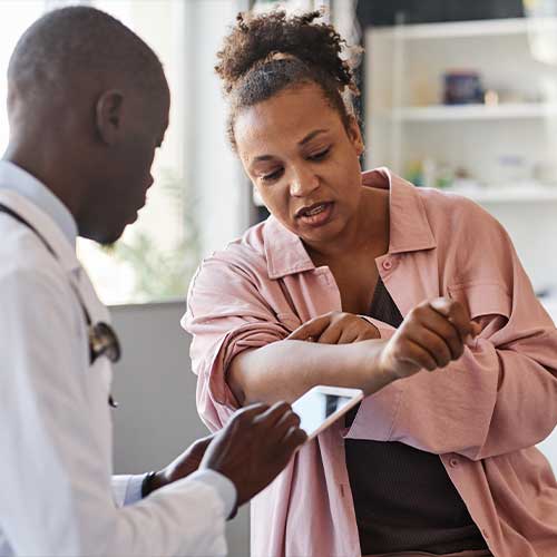Adult African American woman suffering from psoriasis visiting dermatologist in clinic and pointing on rash spots on arms copy space