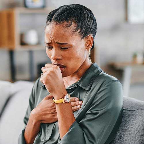 Woman sitting on sofa at home, frowning and pressing on chest, coughing into palm