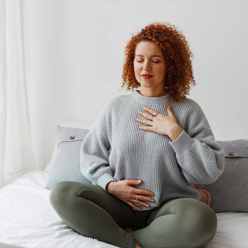 Portrait of plus-size curly redhead female in casual clothes doing pranayama technique sitting on bed next to window with closed eyes, putting hands on chest, breathing slowly and deeply