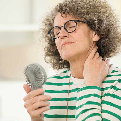 Menopausal mature woman wearing green striped blouse and eyeglasses sitting on sofa in living room at home, having a hot flash and using fan.
