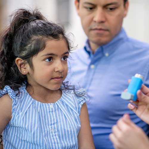 A Father brings his daughter in to the doctor to address his concerns with her Asthma. The female doctor is holding out an inhaler as she reviews the proper administration with the the young girl.