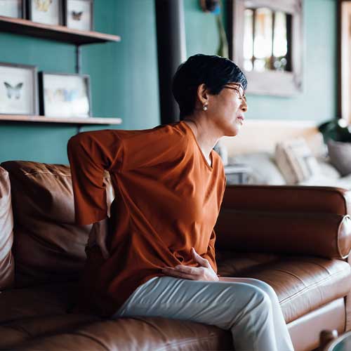 A distraught senior Asian woman feeling unwell, suffering from backache, massaging aching muscles while sitting on sofa in the living room at home. Elderly and health issues concept