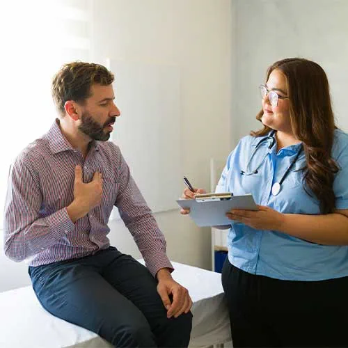 Male patient explaining health concerns while seated on exam table, female physician listening and documenting medical information during consultation