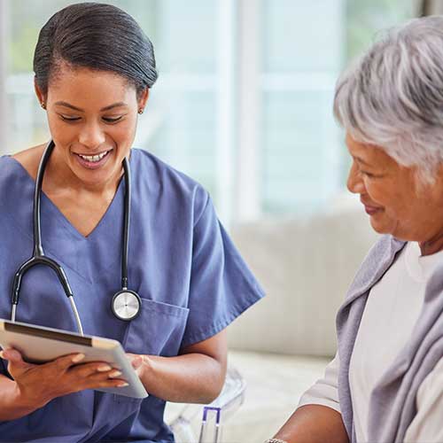 A hispanic senior woman in on a cosy sofa and her female nurse in the old age home and using a digital tablet. Mixed race young nurse and her patient talking in the lounge