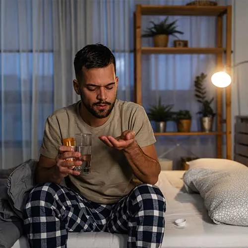 Portrait of a young man holding medicaments in one hand and a glass of water in the other. He is about to drink a recommended dose.