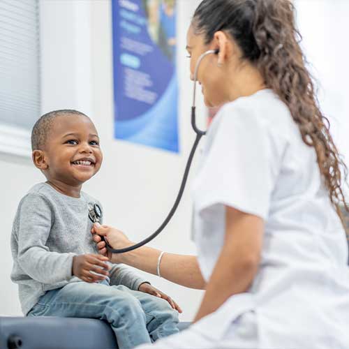 A sweet little boy sits up on an exam table in a doctors office during a routine check-up. His female doctor is seated in front of him and listening to his heart with her stethoscope.