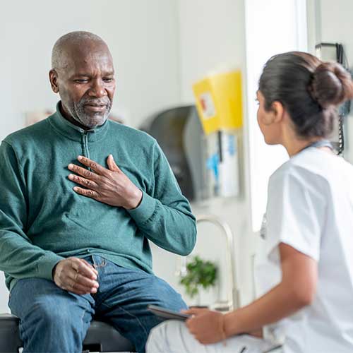 An elderly Black man sits on an exam table, hand on his chest, speaking with a compassionate female nurse in a clinic. A calm, trusting moment highlighting patient care and empathy.