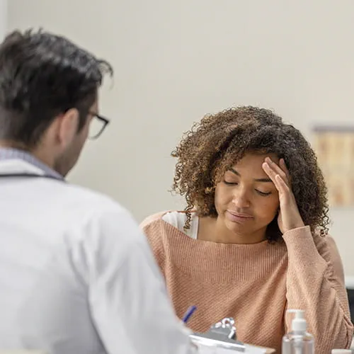 Patient seated across her doctor at a medical appointment with her hand on her forehead