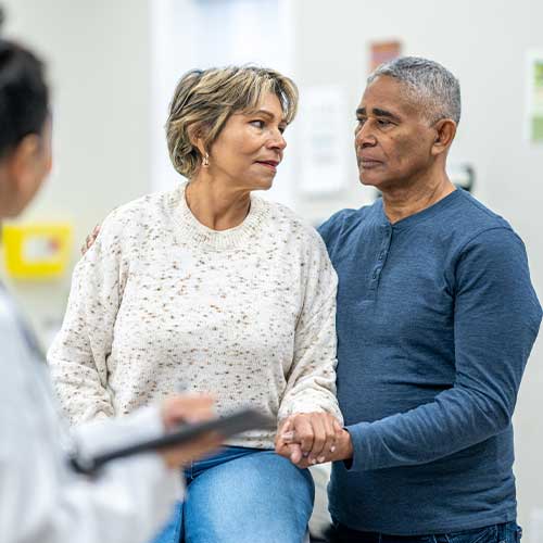 A senior patient, of Hispanic decent, sits up on an exam table with her husband close by as she receives her recent tests results. The couple appears nervous as they talk with their female doctor.