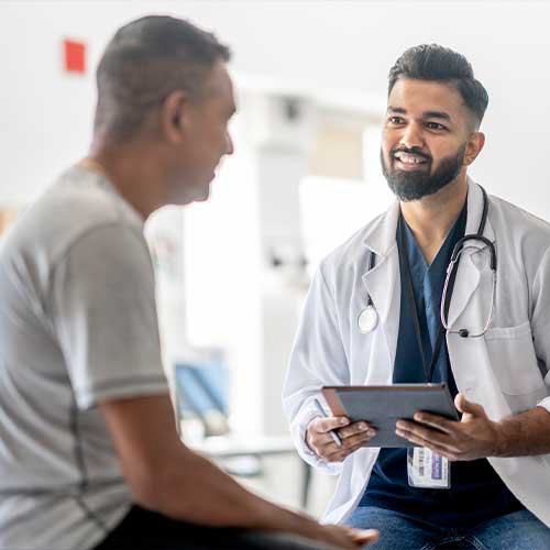 A male doctor of Middle Eastern decent, sits across from his senior male patient as they discuss his health at an appointment. He is wearing a white lab coat and is seated on front of the patient as the senior sits up on the exam table.
