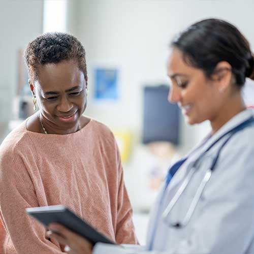 A senior woman of African decent sits up on an exam table during a routine check-up. Her female doctor is holding out a tablet as they review some recent test results together.