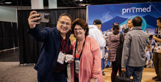 Attendee taking selfie with a colleague with Pri-Med booth and Peter Lin in the background