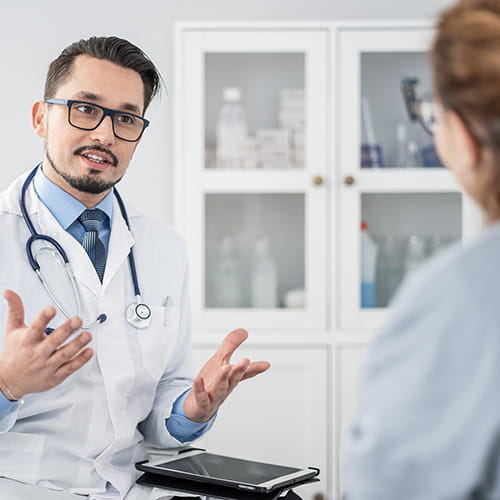 Bearded doctor in glasses, white coat, and stethoscope gesturing as he speaks with a patient in an exam room.