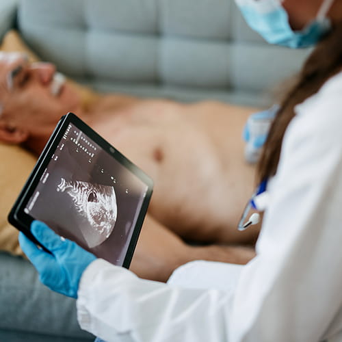 Clinician in gloves and a face mask holds a tablet showing an ultrasound image while a patient lies on a couch in the background.