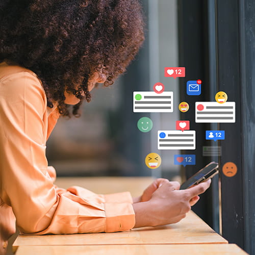 A person scrolling on their phone while social media notifications, likes, and emoji reactions float around them.