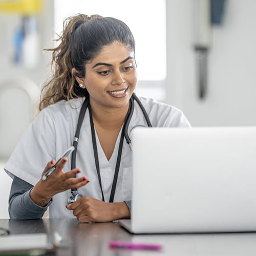 Smiling healthcare provider in scrubs and a stethoscope looking at a laptop during a virtual visit.