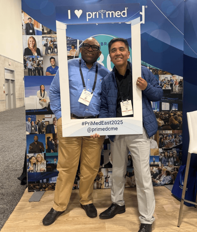 Two attendees at the Pri-Med booth smiling for the camera holding an "I [heart] Pri-Med" sign