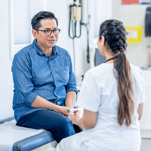 A middle aged man sits on an exam table while talking to his female doctor.