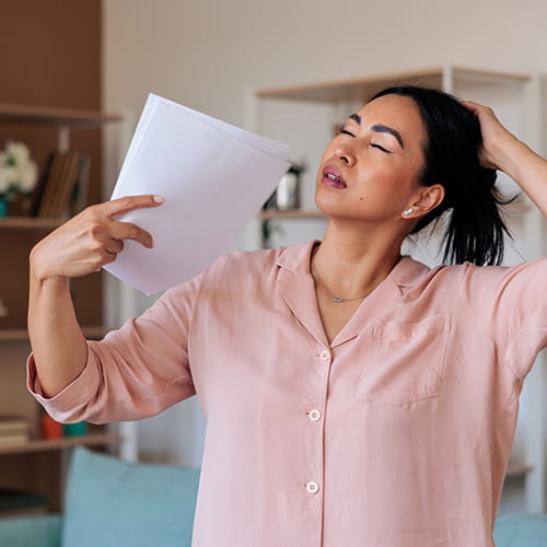 A woman experiencing a hot flash, fanning herself with a paper in one hand while gathering her hair with the other.