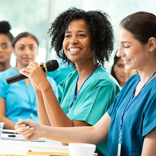 A woman in scrubs speaks confidently to a group of healthcare professionals.