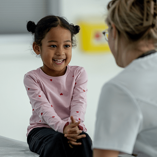 A little girl sits on an exam table in a doctor's office, smiling at her doctor as they talk.