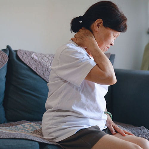 An older Asian woman suffernig from neck pain, rubbing the back of her neck.