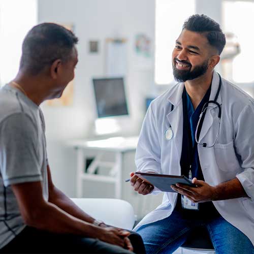 A gentleman of African decent, sits up on an exam table as he talks with his male doctor about his health concerns. The doctor is dressed professionally and taking notes electronically.