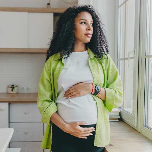 Indoor portrait of african american pensive pregnant woman in green shirt, waiting for her husband to come back from business trip, standing in kitchen looking through window, rubbing cute belly