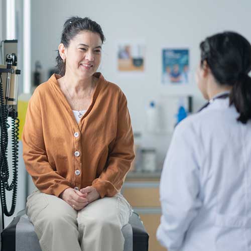 A middle-aged woman of Hispanic decent, sits up on an exam table during a routine check-up. She is dressed casually in an orange sweater and talking with her female doctor about her health.