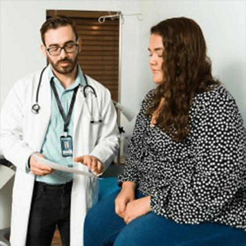 Doctor in a white coat reviewing a document while talking with a seated female patient in an exam room.
